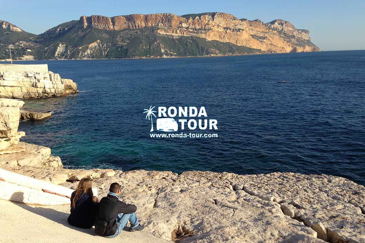 Cap Canaille observé depuis la presqu'ile de Port Miou à Cassis par un couple sur le littoral rocheux. Filigrane avec le logo de Ronda Tour et son adresse web www.ronda-tour.com sur la photo.