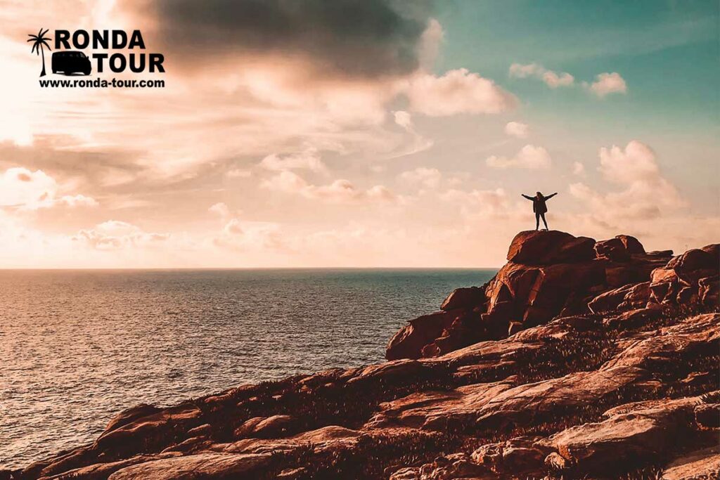 Personne les bras écartés qui regarde la mer du haut des rocher pendant le coucher de soleil. Filigrane avec le logo de Ronda Tour et son adresse web www.ronda-tour.com sur la photo.
